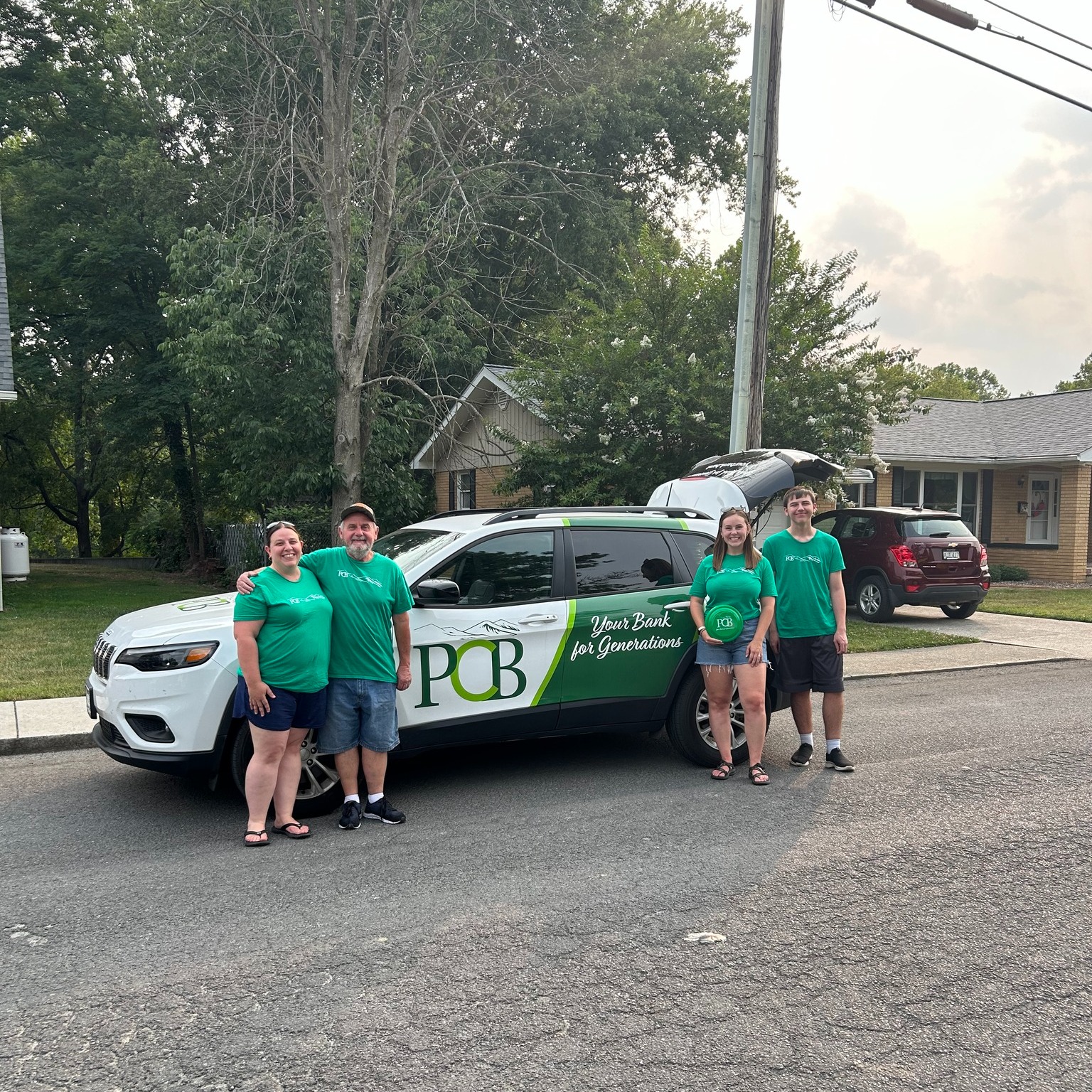 Employees at Petersburg PCB standing next to the PCB branded car in a neighborhood street with trees in the background. 